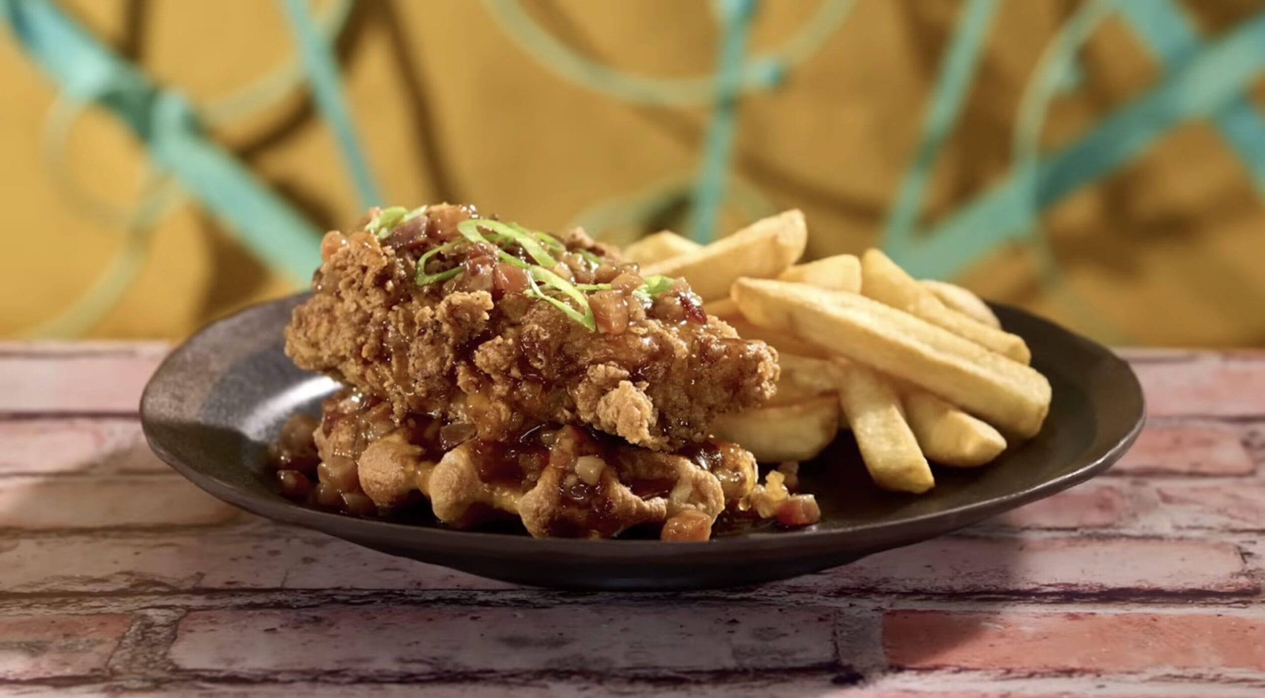 A plate showcasing a delightful fusion dish: fried chicken on waffles, drizzled with sauce and sprinkled with green onions. Accompanied by thick-cut fries, this Food & Beverage delight promises an epic universe of flavors in every bite.